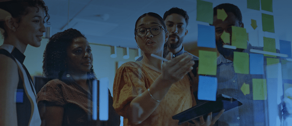 Manager carrying digital tablet, standing in front of glass wall with sticky notes, explaining work strategies to teammates listening attentively in meeting room.